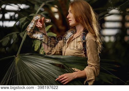 A Beautiful Girl With Natural Make-up And Red Hair Stands In The Jungle Among Exotic Plants With Sna