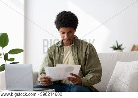 Happy Black Guy Learning Online From Home, Writing Coursework, Holding Documents Near Laptop. Africa