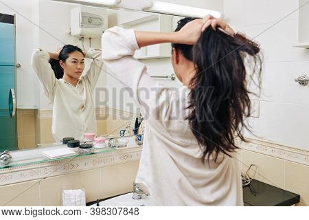 Pretty Young Chienese Woman In White Shirt Making High Ponytail In Front Of Mirror In Bathroom
