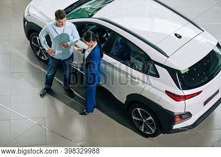 Dealership Manager Explaining Payment Details To Customer When Thay Are Standing At Automobile, View