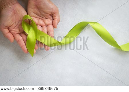 Hands Holding Light Lime Green Ribbon On White Fabric Background With Copy Space. World Mental Healt