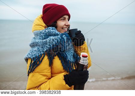 Young Woman Pours Hot Tea From A Thermos Into A Cup And Drinks On The Seaside. Female Tourist Is Hav