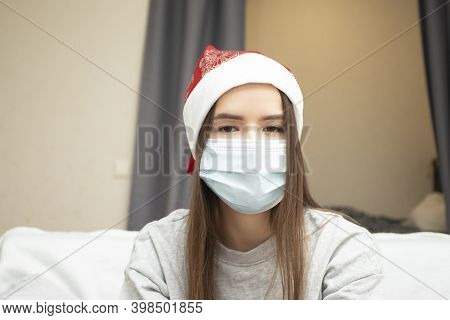 Coronavirus Christmas 2021. Portrait Of A Young Woman In A Medical Mask And A New Yerar Hat At Home