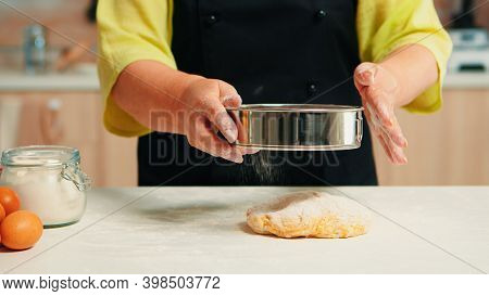 Senior Woman With Black Apron Sifting Flour On Dough Using Metallic Sieve Happy Elderly Chef With Bo