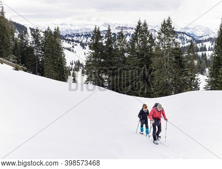 Senior Couple Is Snowshoe Hiking In Alpine Snow Winter Mountains. Allgau, Bavaria, Germany.