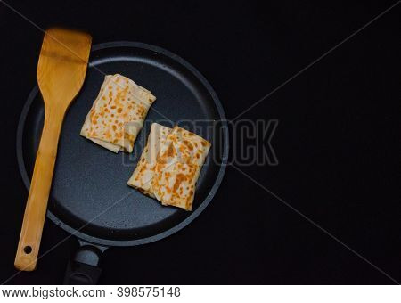 Thin Pancakes With Filling On A Black Frying Pan And A Black Background With A Wooden Spatula