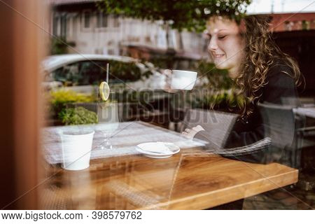 Cute Brunette, Curly Girl Sitting In A Cafe, Drinking Coffee And Juice. View Though The Wondow