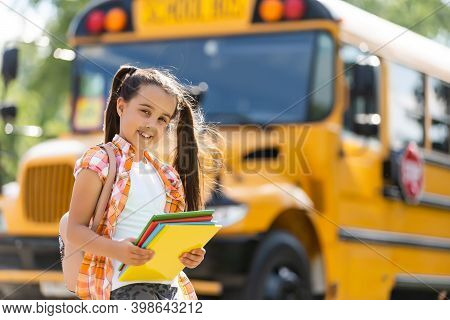 Adorable Little Schoolgirl Near School Bus And Looking At Camera
