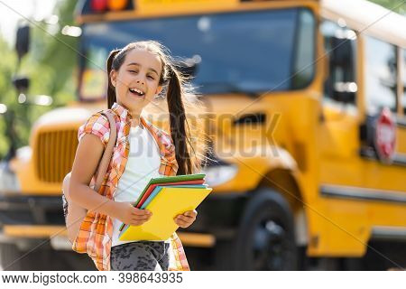 Adorable Little Schoolgirl Near School Bus And Looking At Camera