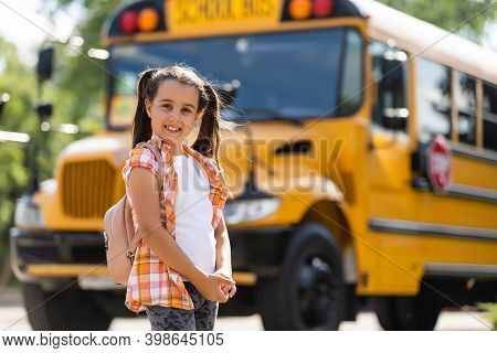 Adorable Little Schoolgirl Near School Bus And Looking At Camera