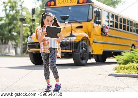 Adorable Little Schoolgirl Near School Bus And Looking At Camera