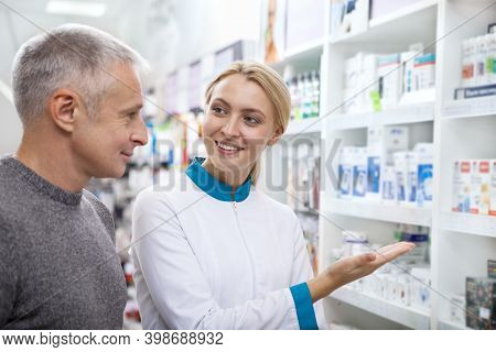 Friendly Female Chemist Helping Her Senior Male Customer, Showing Pharmacy Assortment On The Shelves