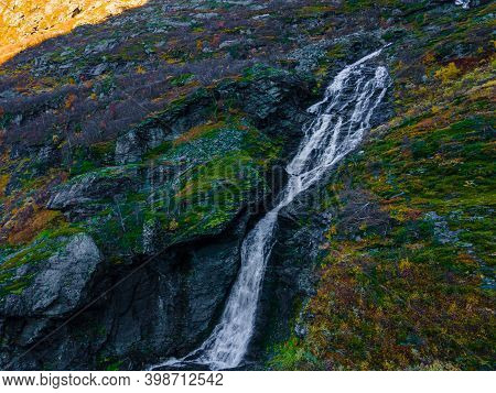 Large Waterfall On A Mountain Side With Green Turf And Large Boulders.