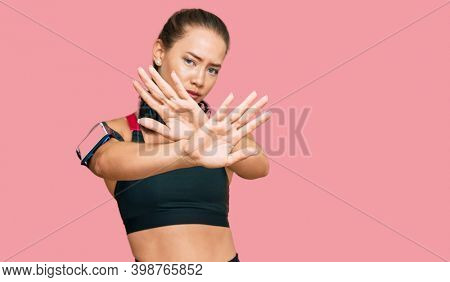 Beautiful blonde woman wearing gym clothes and using headphones rejection expression crossing arms and palms doing negative sign, angry face 