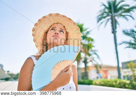 Young beautiful caucasian woman with blond hair smiling happy outdoors using handfan on a hot summer day