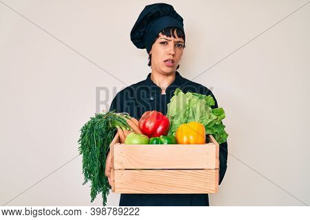 Beautiful brunettte woman chef holding fresh veggetables clueless and confused expression. doubt concept. 