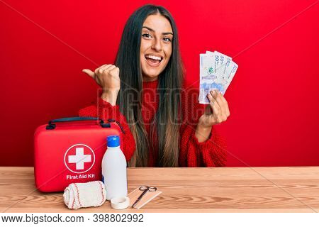Beautiful hispanic woman with first aid kit holding colombian pesos pointing thumb up to the side smiling happy with open mouth 