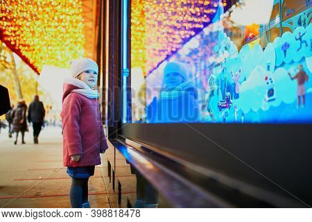 Toddler Girl Looking At Window Glass Of Large Department Store Decorated For Christmas. Child Enjoyi