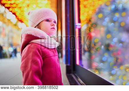 Toddler Girl Looking At Window Glass Of Large Department Store Decorated For Christmas. Child Enjoyi
