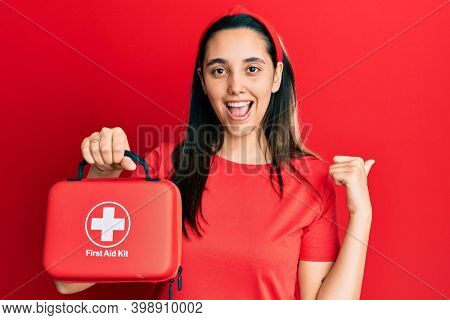 Young hispanic woman holding first aid kit pointing thumb up to the side smiling happy with open mouth 