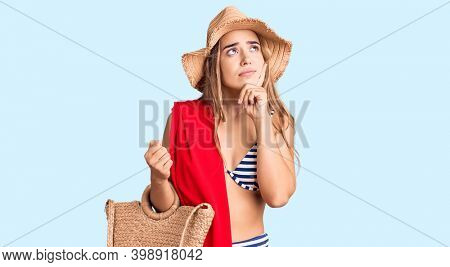 Young beautiful blonde woman wearing bikini and hat holding summer wicker handbag serious face thinking about question with hand on chin, thoughtful about confusing idea 