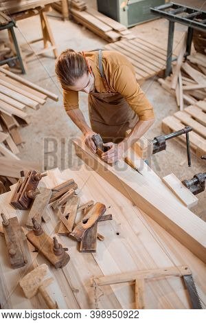 Handsome Carpenter Working With A Wood, Planing A Bar With A Plane On The Worbench In The Carpentry 