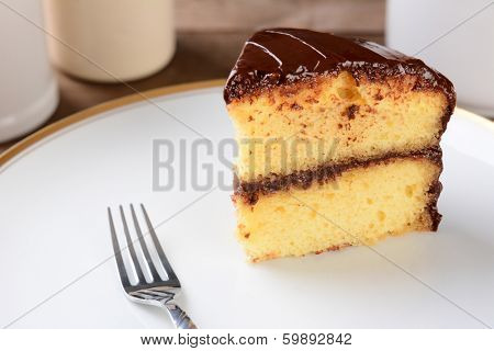 A slice of yellow cake with chocolate frosting on a white plate with a fork.