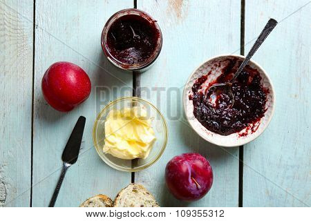 Tasty jam in the jar and bowl, butter, fresh bread, plums and tablet on blue wooden background