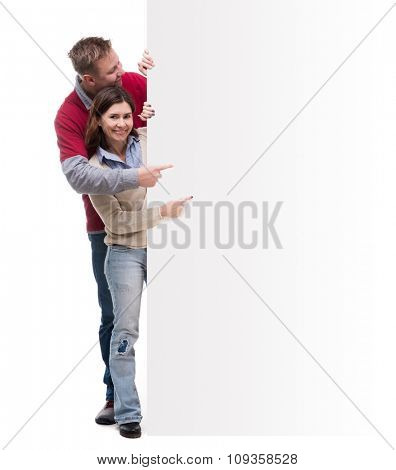 cute cuople peek out from behind the blank pointing at empty space isolated on white background