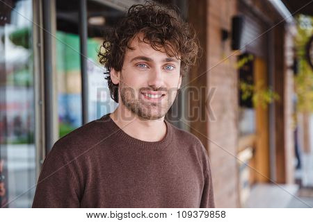 Portrait of happy handsome positive attractive curly smiling young man in brown sweetshirt in the city