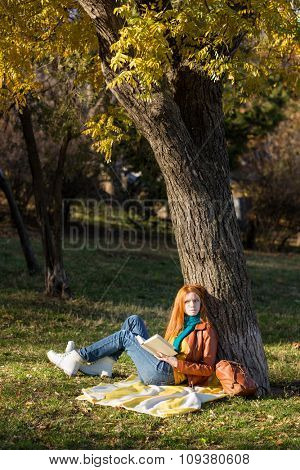 Thoughtful young redhead woman reading a book sitting under the tree in park and dreaming