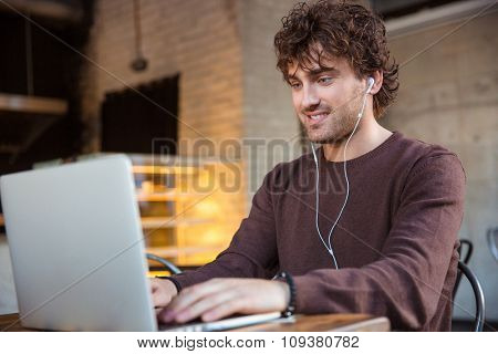 Concentrated smiling happy pleased content curly male in brown sweetshirt using laptop and listening to music
