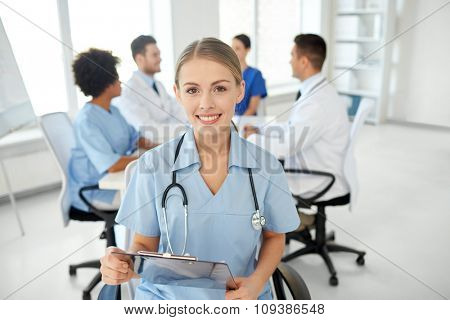 health care, profession, people and medicine concept - happy female doctor or nurse with clipboard over group of medics meeting at hospital