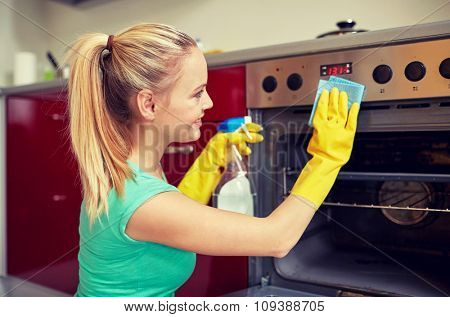 people, housework and housekeeping concept - happy woman with bottle of spray cleanser cleaning oven at home kitchen