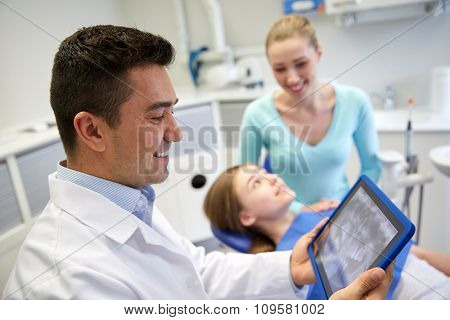 people, medicine, stomatology, technology and health care concept - happy male dentist looking at teeth x-ray on tablet pc computer and patient girl with her mother at dental clinic office