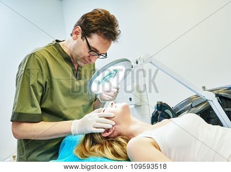 Male doctor cosmetologist examining young woman skin with loupe before the cosmetology procedure