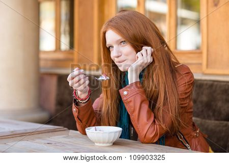 Cute content young woman with beautiful long red hair having breakfast in outdoor cafe and talking on cellphone biting bottom lip