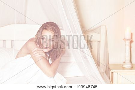 Portrait of gentle romantic woman sitting in the bed under gentle transparent valance, spending weekend in luxury spa hotel, enjoying rest and relaxation