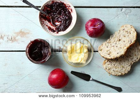 Tasty jam in the jar and bowl, butter, fresh bread, plums and tablet on blue wooden background