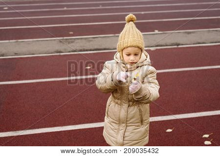 Little 4 year-old girl in a beige woolen hat and warm holding a little yellow flower on a race track.