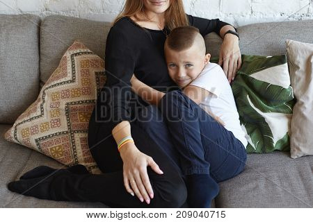 Sissy mama's boy sitting on grey couch and hugging his unrecognizable mother seeking for her support against bullies not able to stand up for himself having miserable expression on his face