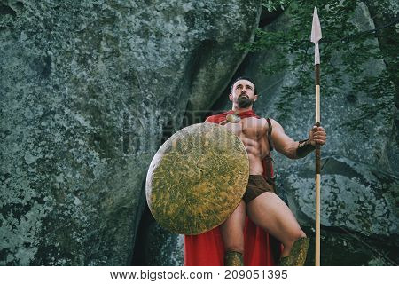 Low angle shot of a strong brave muscular Spartan warrior holding a spear and a shield posing in the woods copyspace.