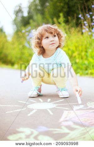 Sunny and warm summer days and happy chilhood. Cute little girl drawing with chalk crayons at the asphalt. Child Having fun alone. Spending time without parents and adults. Kids drawings: sun house flowers.