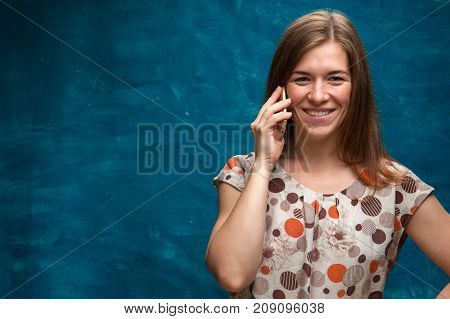 Happy cheerful female looking at the camera and talking on the mobile phone. Portrait of beautiful smiling caucasian woman using her smartphone. Blue backdrop studio background. Copy Space.