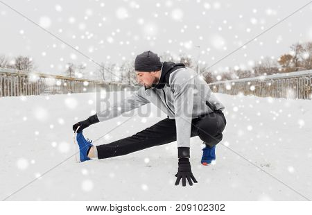 fitness, sport, people, exercising and healthy lifestyle concept - young man stretching leg and warmig up on snow covered winter bridge
