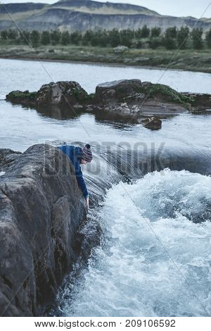 a woman tourist in waterproof clothes and red hat stands on the Bank of the mountain river with small waterfall. Glanni waterfall, Iceland