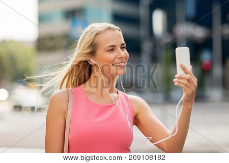 technology, lifestyle and people concept - smiling young woman with smartphone and earphones listening to music and taking selfie in city