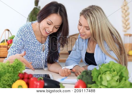 Two young happy women are making online shopping by tablet computer and credit card. Friends are going to cook in the kitchen, they searching internet for a new recipe