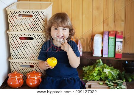 Adorable happy toddler girl sitting in the kithcen idoors eating yellow tomato smiling.