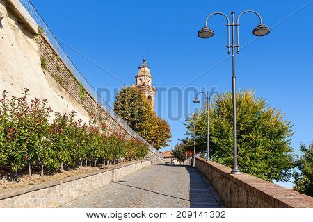 Narrow street leading towards belfry along lampposts and autumnal trees under blue sky in small town of Diano d'Alba in Piedmont, Northern Italy.